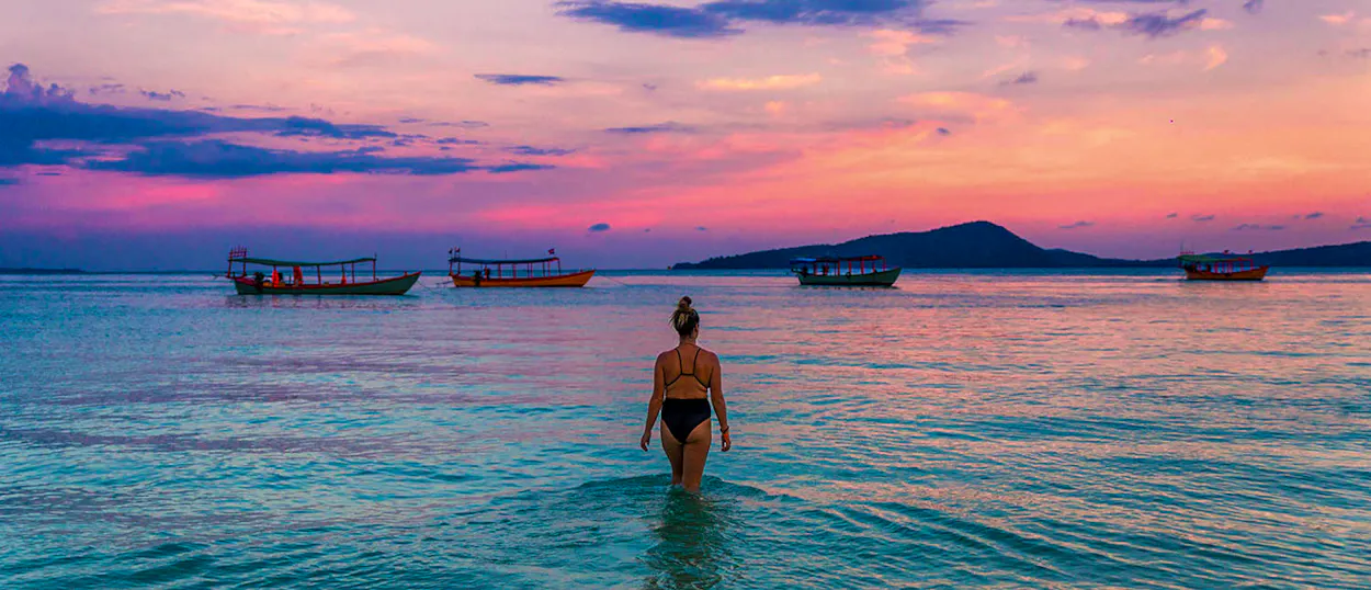 A woman wades into the clear turquoise waters of Koh Rong at sunset, with traditional boats.