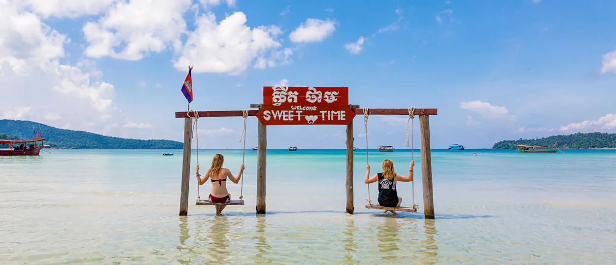 Two women enjoy swings in the clear turquoise waters of Koh Rong, Cambodia, under a sunny sky.