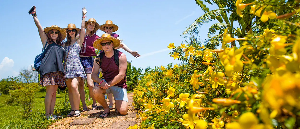 Happy tourists in straw hats pose on a sunny path with vibrant yellow flowers in Kampot.
