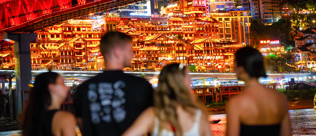 Travellers look across at a brightly lit cliffside building beneath a red steel bridge at night.