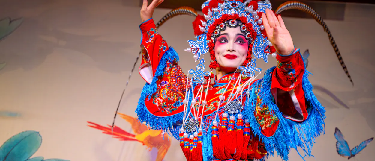 A Sichuan Opera performer in a painted mask and red embroidered costume strikes a pose on stage.