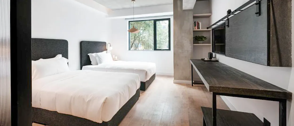 A minimalist twin room with dark headboards, concrete walls and a window onto trees.