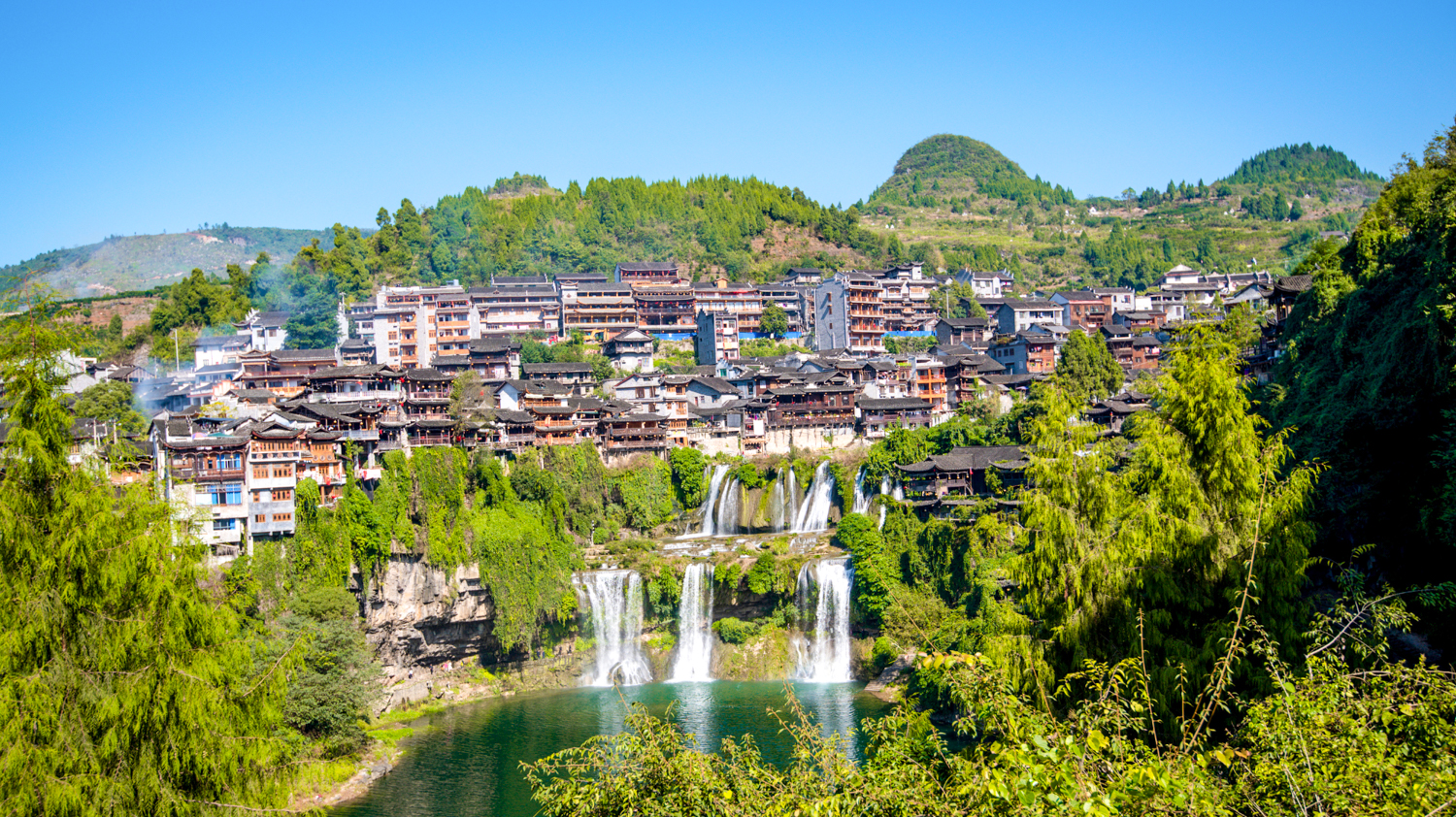 Traditional buildings of Furong Ancient Town, China, perched above a beautiful waterfall and lush green landscape.