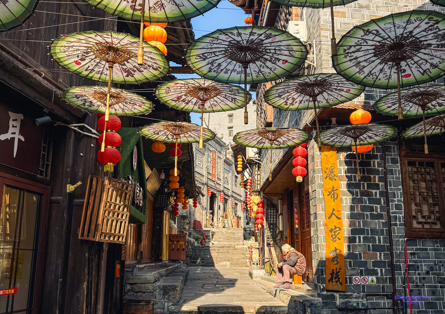 Traditional umbrellas and red lanterns adorn a sunny, stepped street in Furong Ancient Town, with a person.