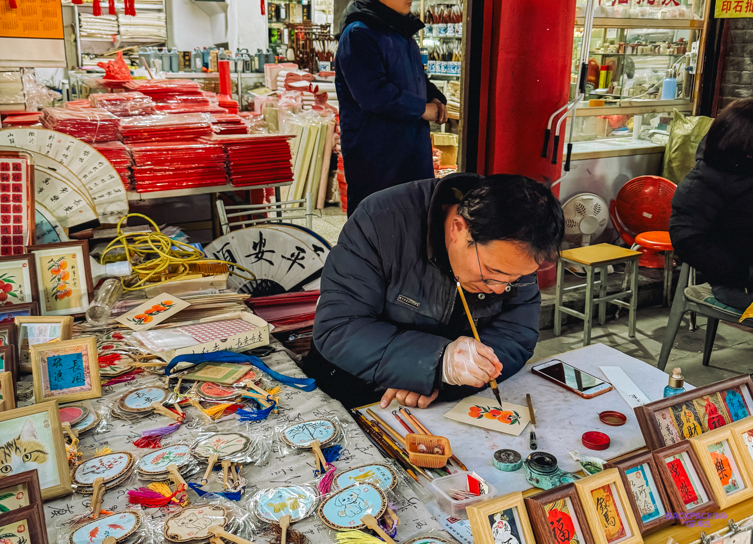 An artist meticulously paints small crafts at a bustling evening market stall in Xi'an, China.