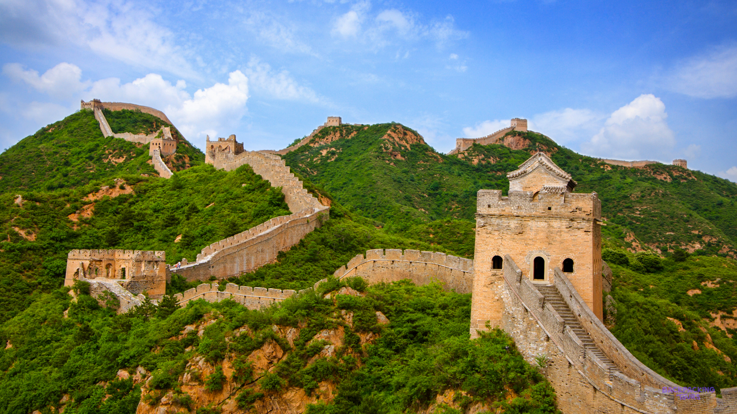The Great Wall of China snakes across verdant mountains with ancient watchtowers under a bright blue sky.
