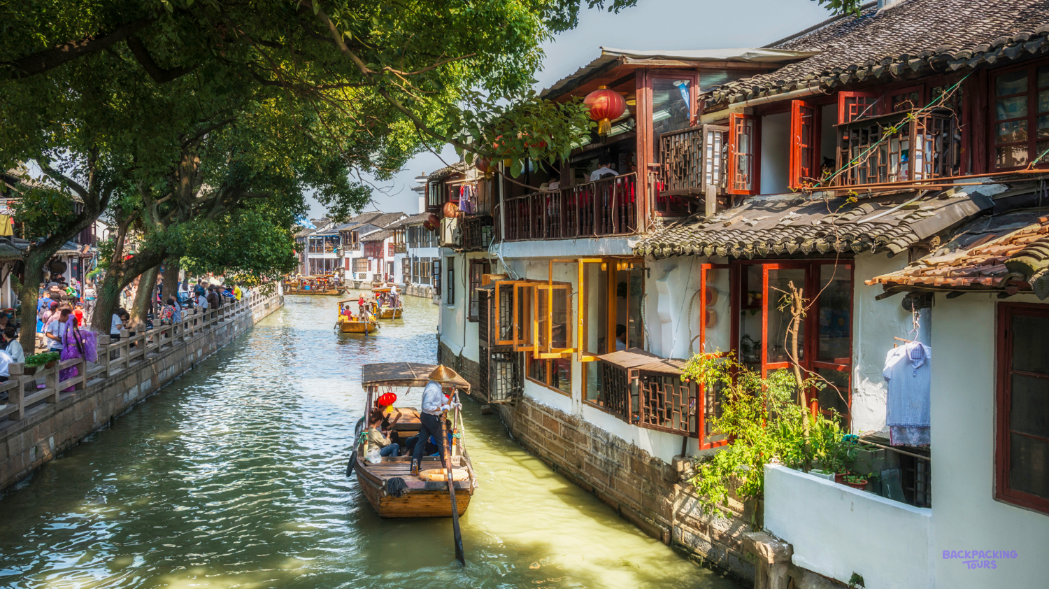 Traditional boats navigate a bustling canal flanked by historic buildings and lush trees in Shanghai.
