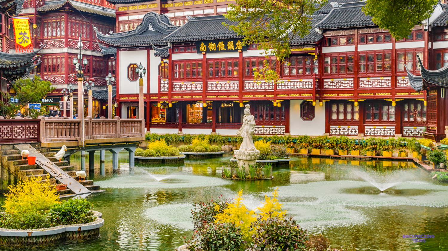 Traditional red Chinese buildings overlook a tranquil pond with fountains and yellow flowers in Shanghai.