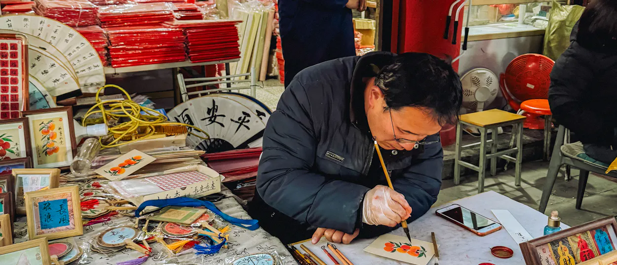 A man meticulously paints small floral designs on crafts at a bustling market in Xi'an.