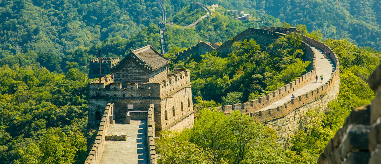 Tourists walk along the ancient Great Wall of China, winding through lush green mountains.