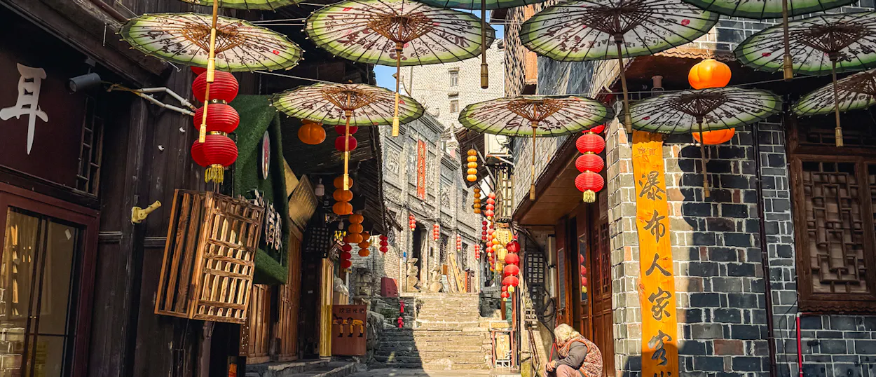 A person sits on a narrow, uphill street in Furong Ancient Town, decorated with lanterns and umbrellas.