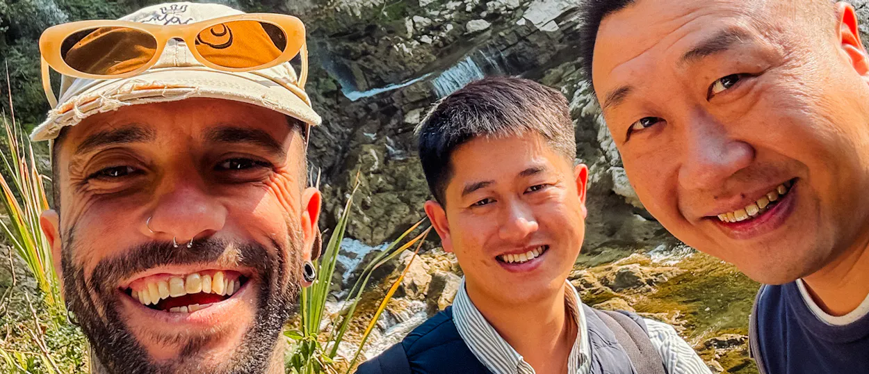Three smiling men take a selfie with a rocky waterfall and greenery in Furong Ancient Town.