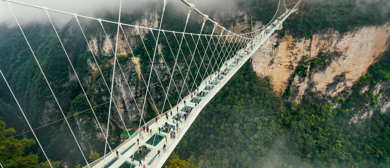 People walk across the Zhangjiajie Glass Bridge, suspended high above a misty, forested valley.