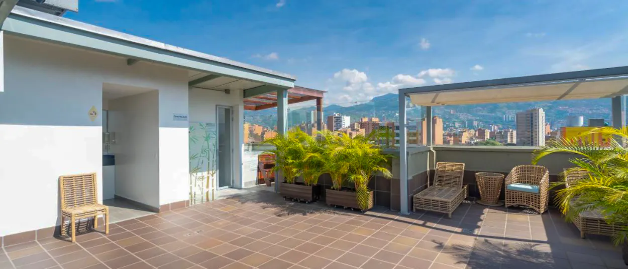 Sunny rooftop terrace with wicker chairs, potted palms, and a panoramic view of Medellin city.