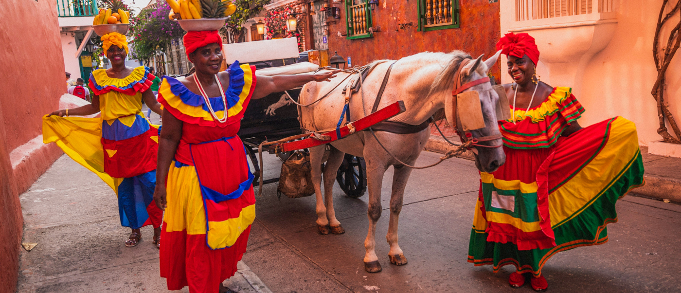 Three women in vibrant traditional dresses with fruit baskets and a horse-drawn carriage in Cartagena.