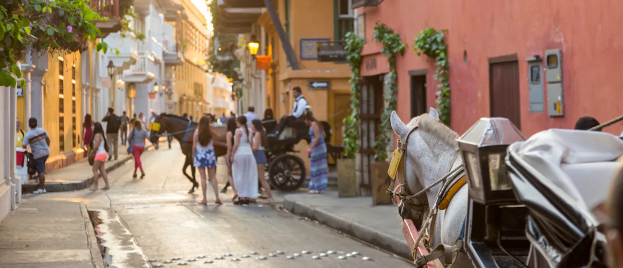 People stroll past colorful buildings and horse-drawn carriages on a vibrant street in Cartagena.