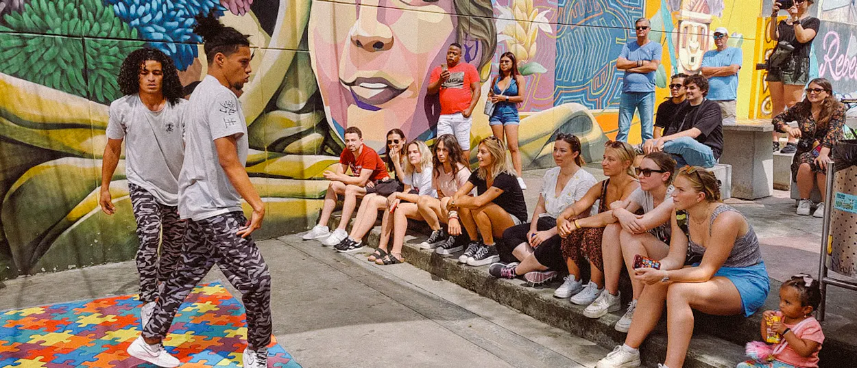 Two men perform street dance in front of a vibrant mural as tourists watch in Medellín.