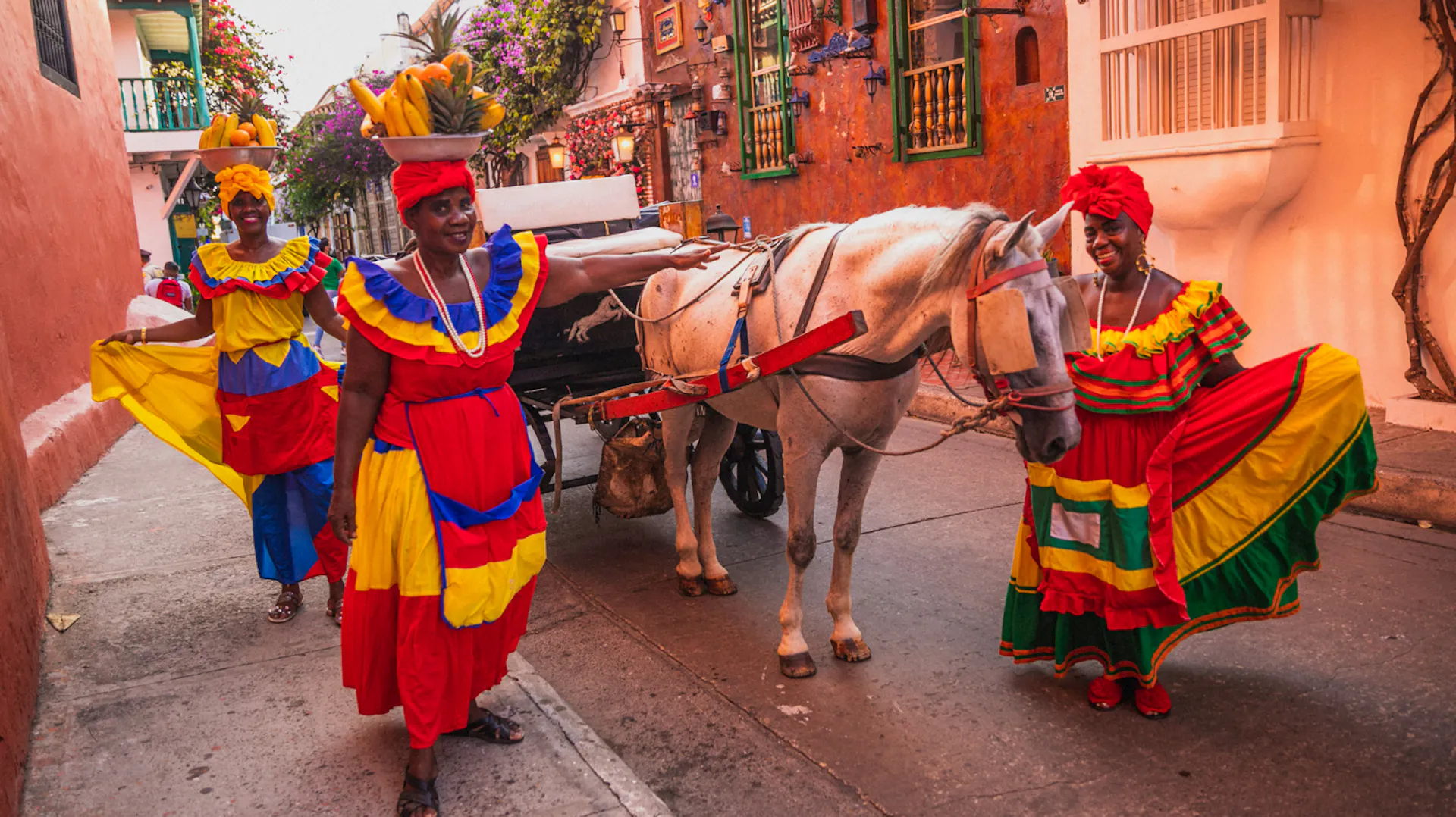 Three smiling women in vibrant Colombian dresses pose with a horse carriage on a colorful street in Cartagena.