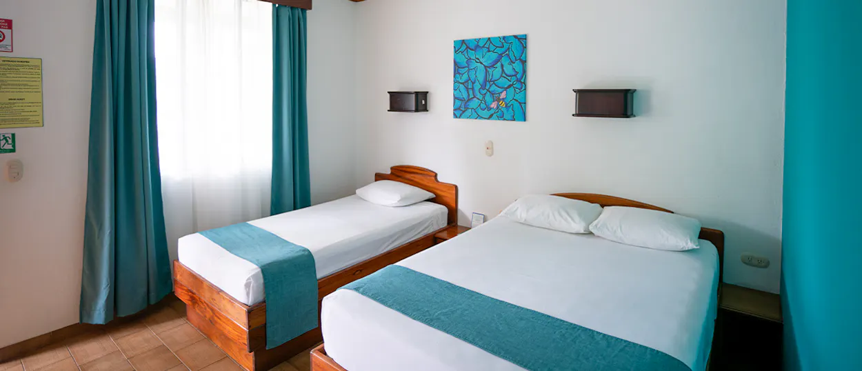 A bright, clean hotel room with a single and a double bed, featuring teal bed runners and curtains.