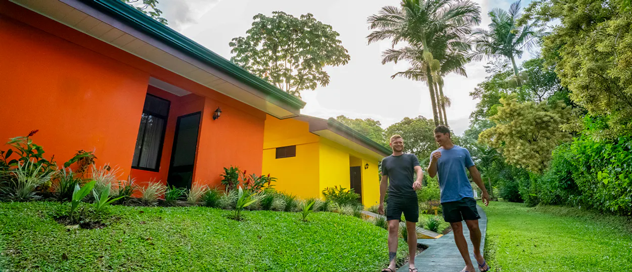 Two men walk past vibrant orange and yellow buildings amidst lush tropical plants in La Fortuna, Costa Rica.