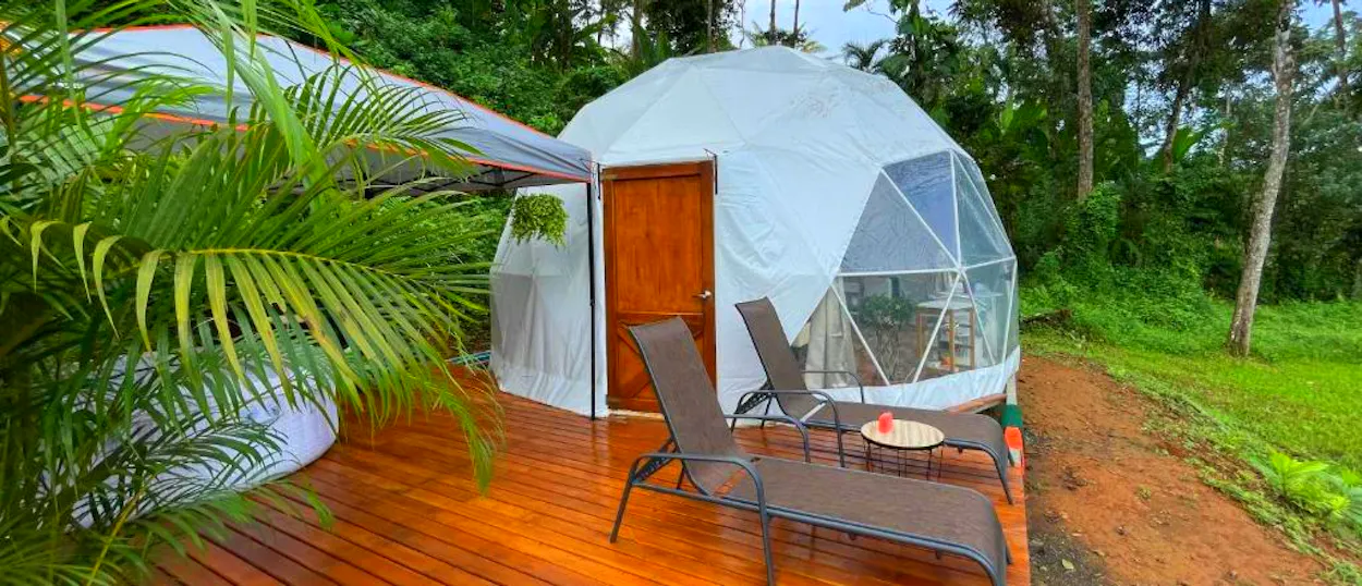 A white glamping dome on a wooden deck with lounge chairs, surrounded by lush tropical greenery.