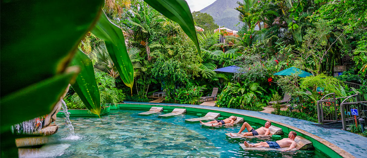 Several men float on loungers in a clear hot spring pool amidst dense Costa Rican jungle.