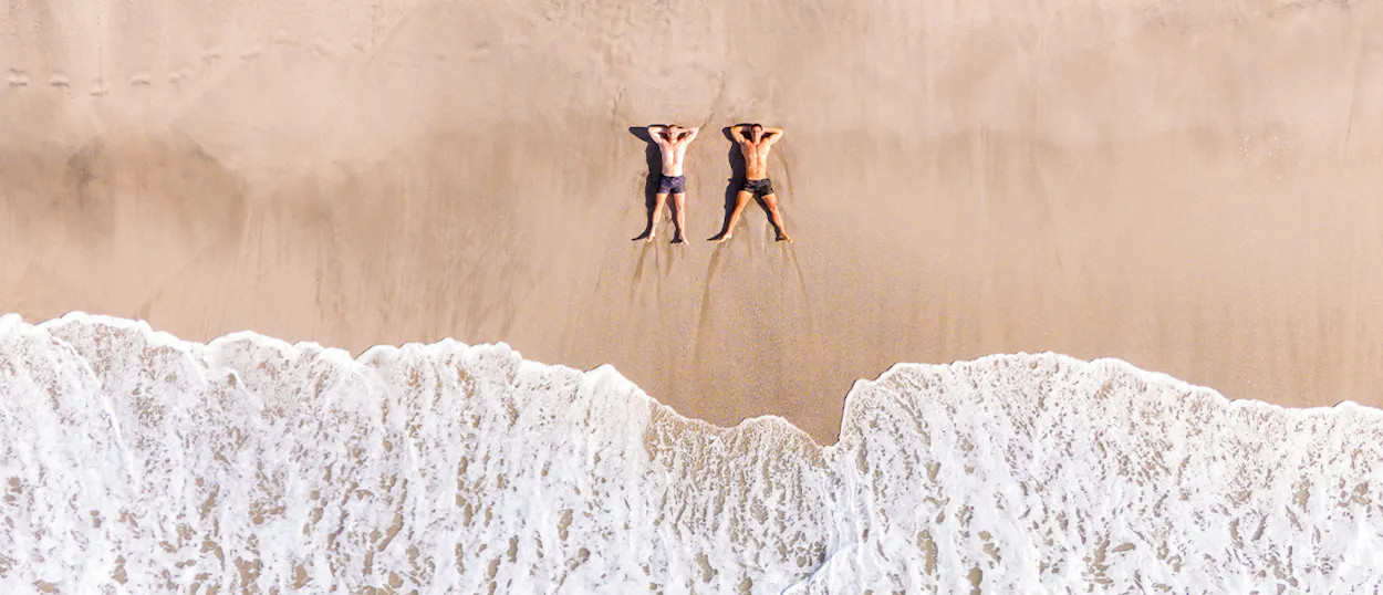 Two men lie relaxed on a golden sandy beach, hands behind heads, as ocean waves roll in.