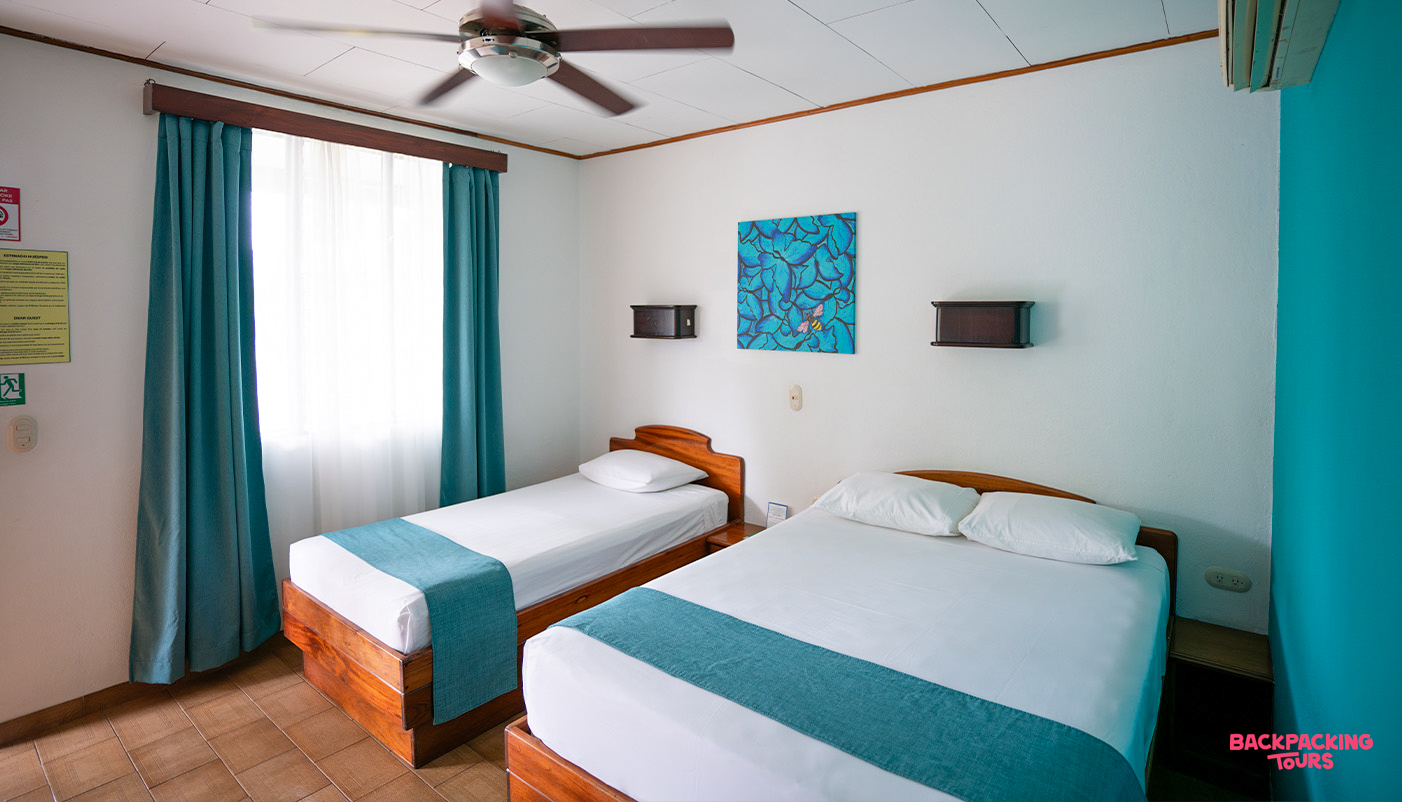 A bright and clean hotel room with two comfortable beds, blue accents, and a ceiling fan.