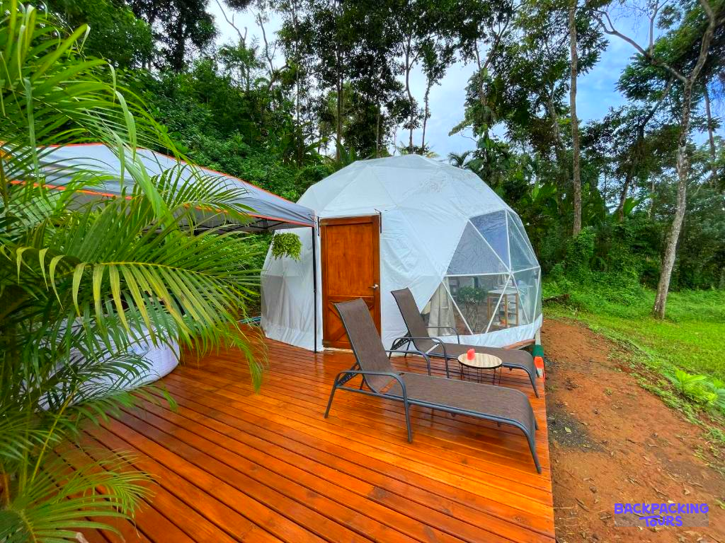 A modern white glamping dome with a wooden deck and lounge chairs in a lush Costa Rican setting.