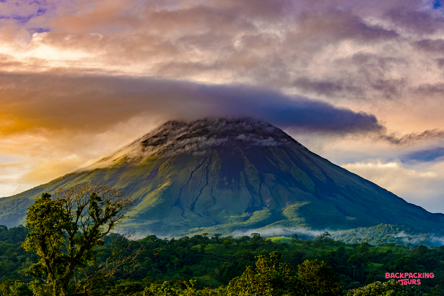 Majestic Arenal Volcano rises above lush rainforest, its peak crowned by dramatic, colorful clouds.