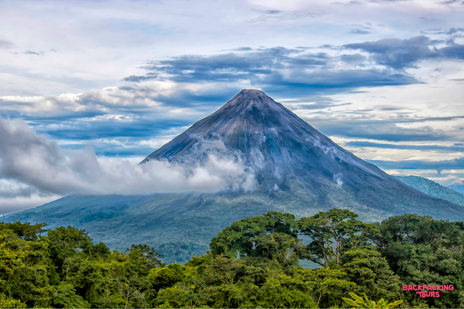 Majestic Arenal Volcano in Costa Rica, with its peak emerging from swirling clouds above lush forest.