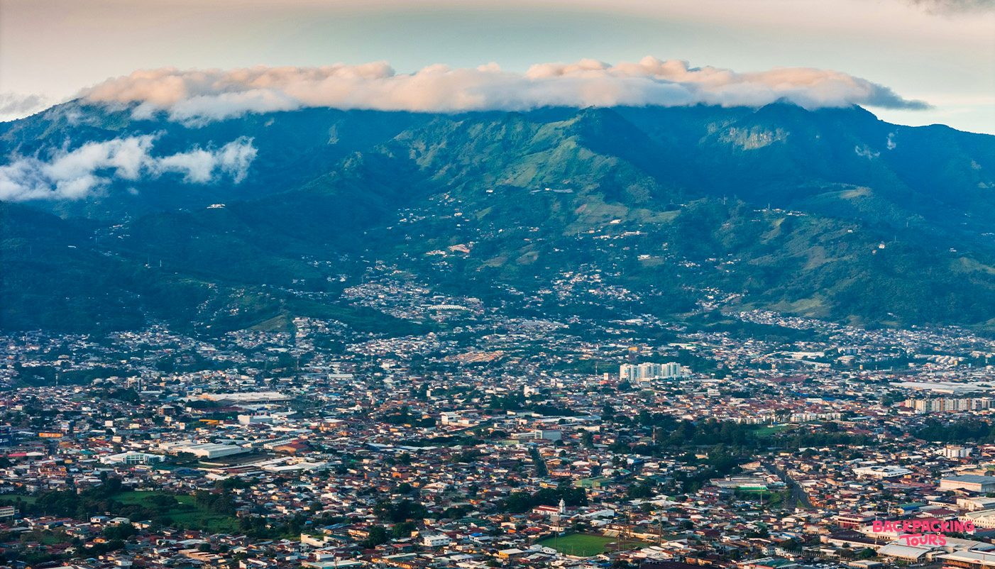 An aerial view of San Jos es, Costa Rica, nestled at the base of lush green mountains with clouds.