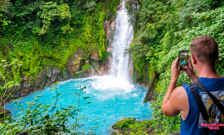 A visitor photographs the vibrant blue waters of a stunning waterfall in the Costa Rican rainforest.