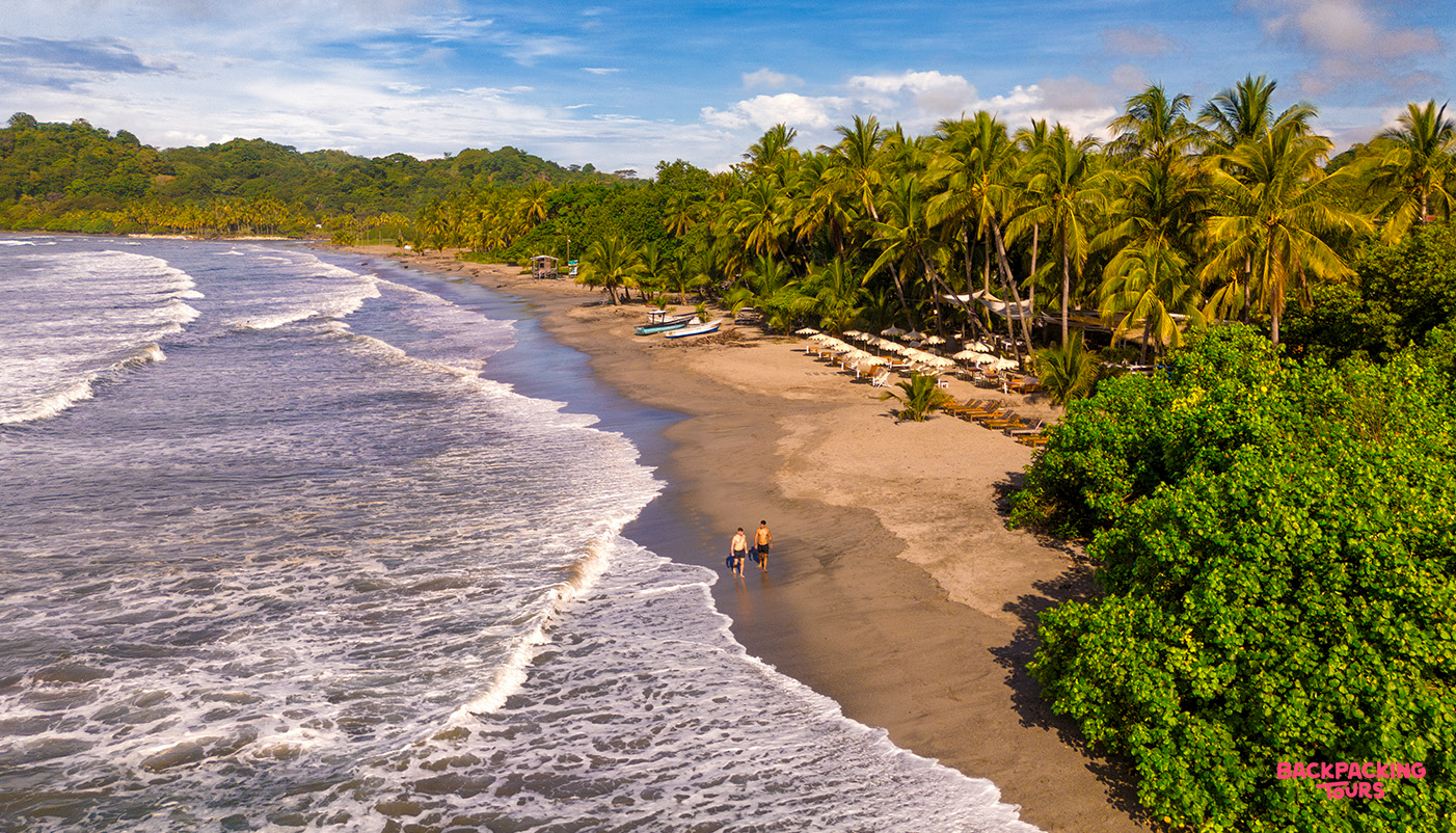Two people walk along the dark sand beach in Samara, Costa Rica, with waves and palm trees.