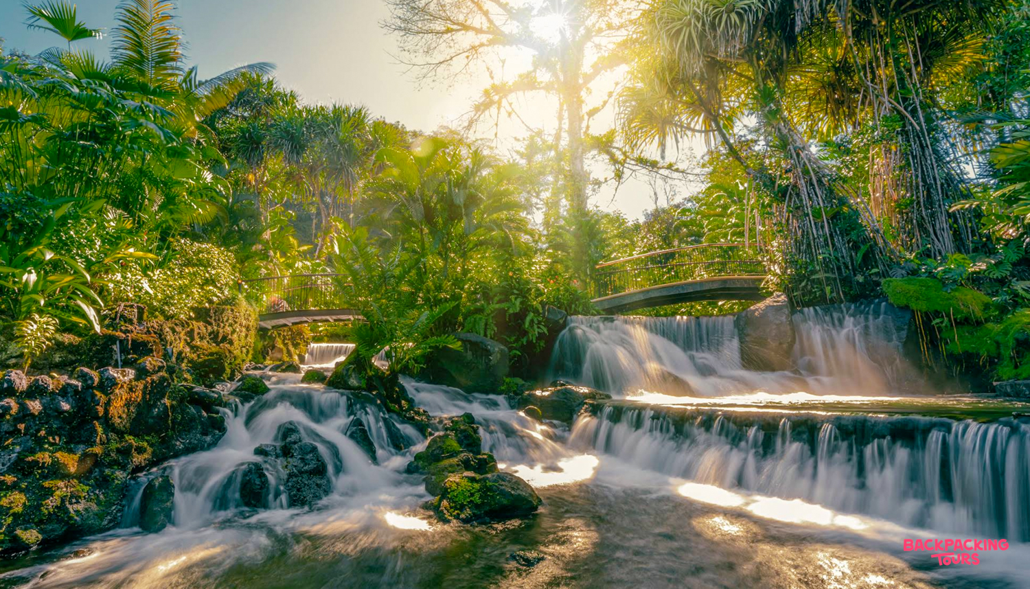 Sunlight illuminates tiered waterfalls and natural hot spring pools nestled within a vibrant tropical rainforest.