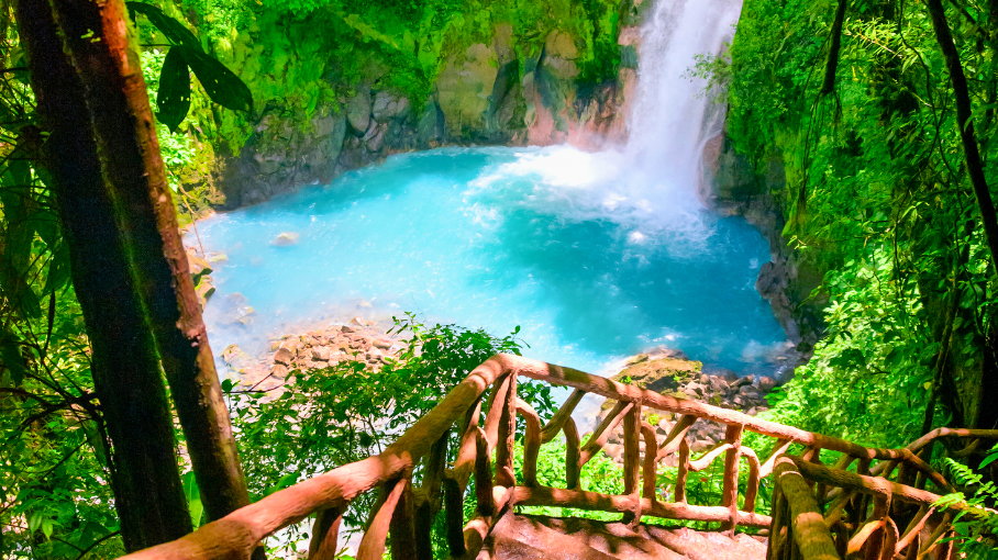 Rustic railing overlooks a vibrant blue waterfall and pool in Costa Rica's lush jungle.