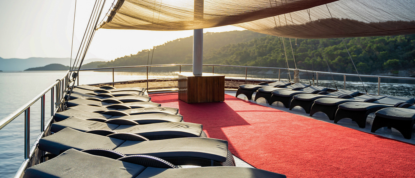 Relaxing sun deck of a sailing boat with lounge chairs and red carpet, sailing past green hills.