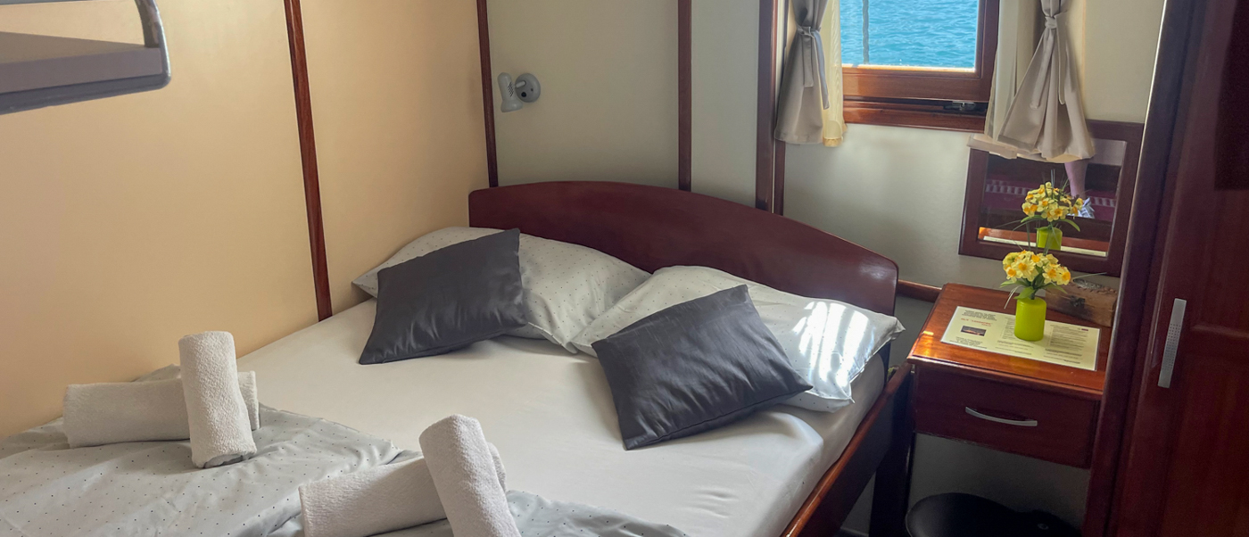 A neat boat cabin with a bed, grey pillows, rolled towels, and a window showing blue water.