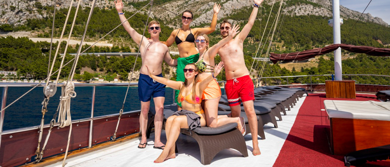 Six happy friends in swimwear wave on a boat deck with a scenic Croatian coastline.