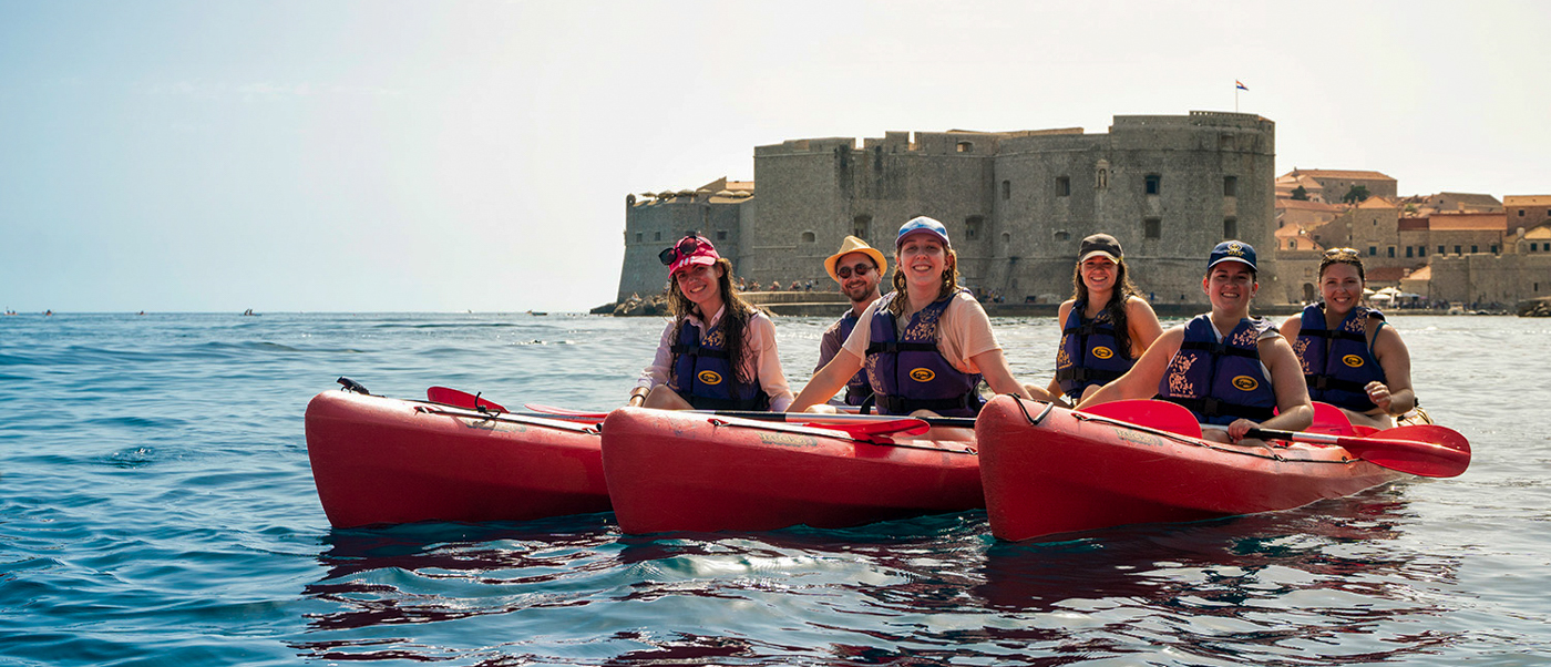 Happy travelers kayak on the clear blue sea, exploring Dubrovnik's ancient fortress walls under a sunny sky.