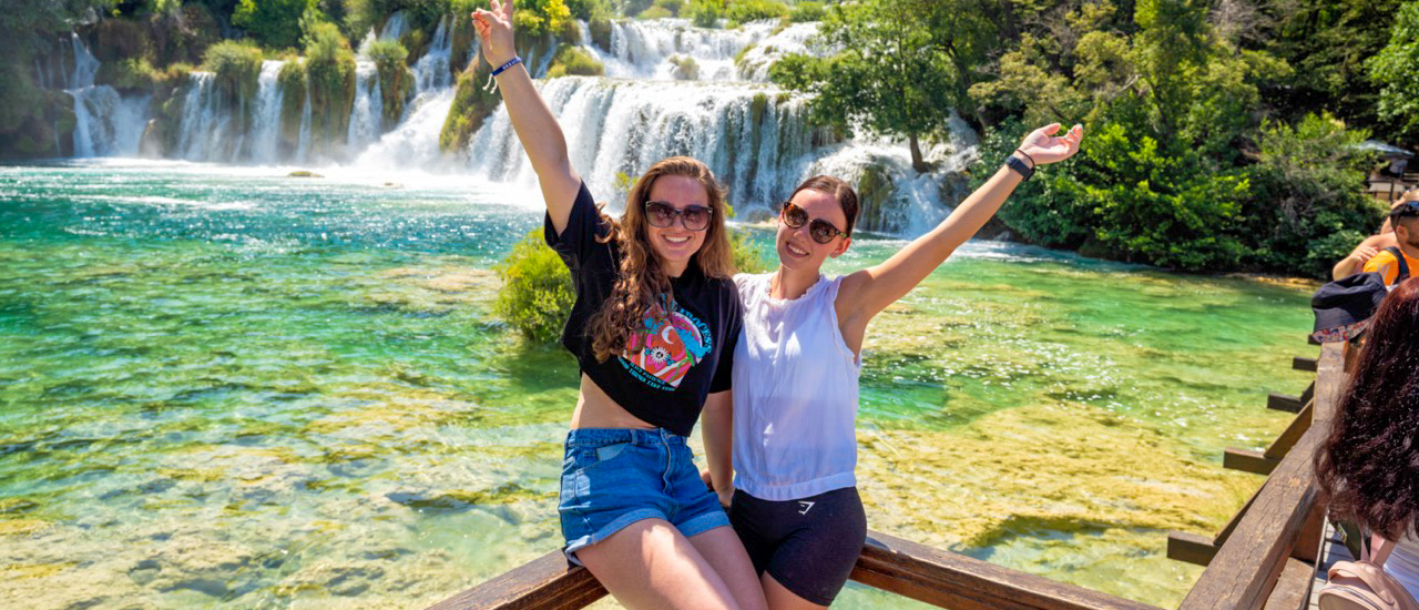 Two happy women with arms raised, smiling in front of the stunning Krka Waterfalls.
