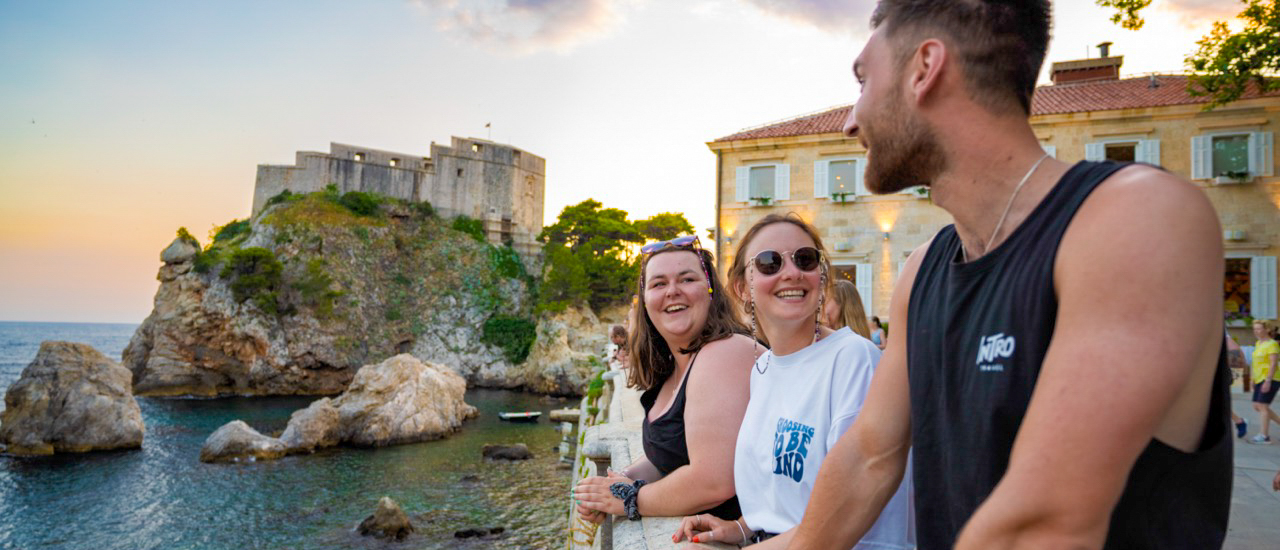 Smiling friends watch the sunset over the historic Dubrovnik fortress and Adriatic Sea.