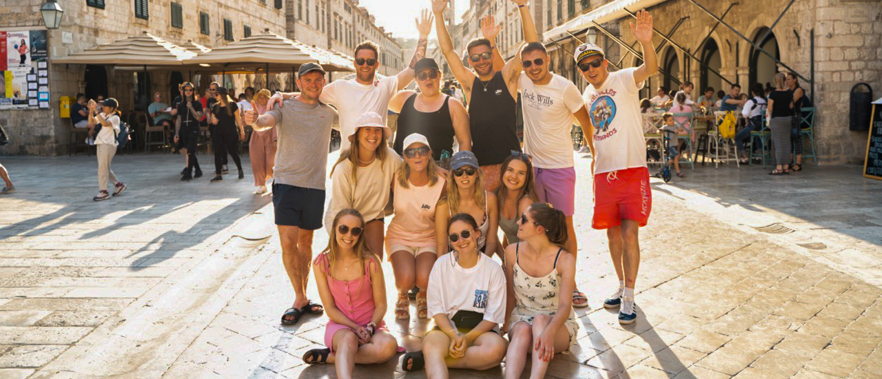 A happy group of backpackers waves to the camera on a sunny, cobbled street in Dubrovnik.