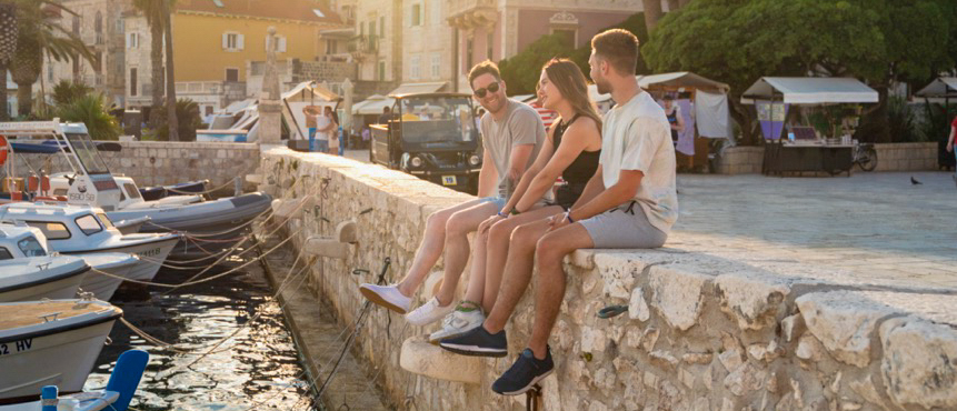 Three friends enjoy a golden sunset from a stone wall overlooking boats in Hvar.