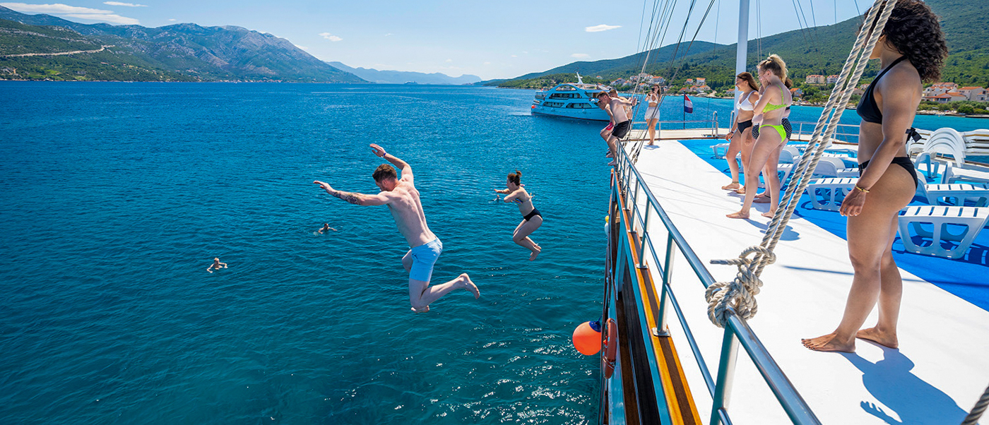 Young people joyfully jump from a boat into the clear blue sea under a sunny sky in Trstenik.
