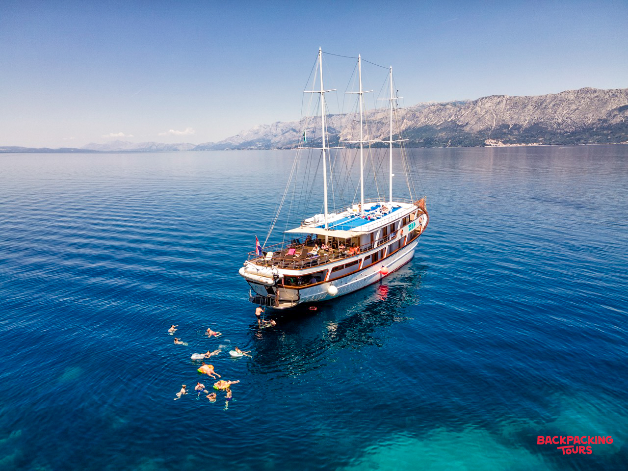 Backpackers swim in the crystal-clear blue waters of Trstenik, Croatia, beside their sailing ship.
