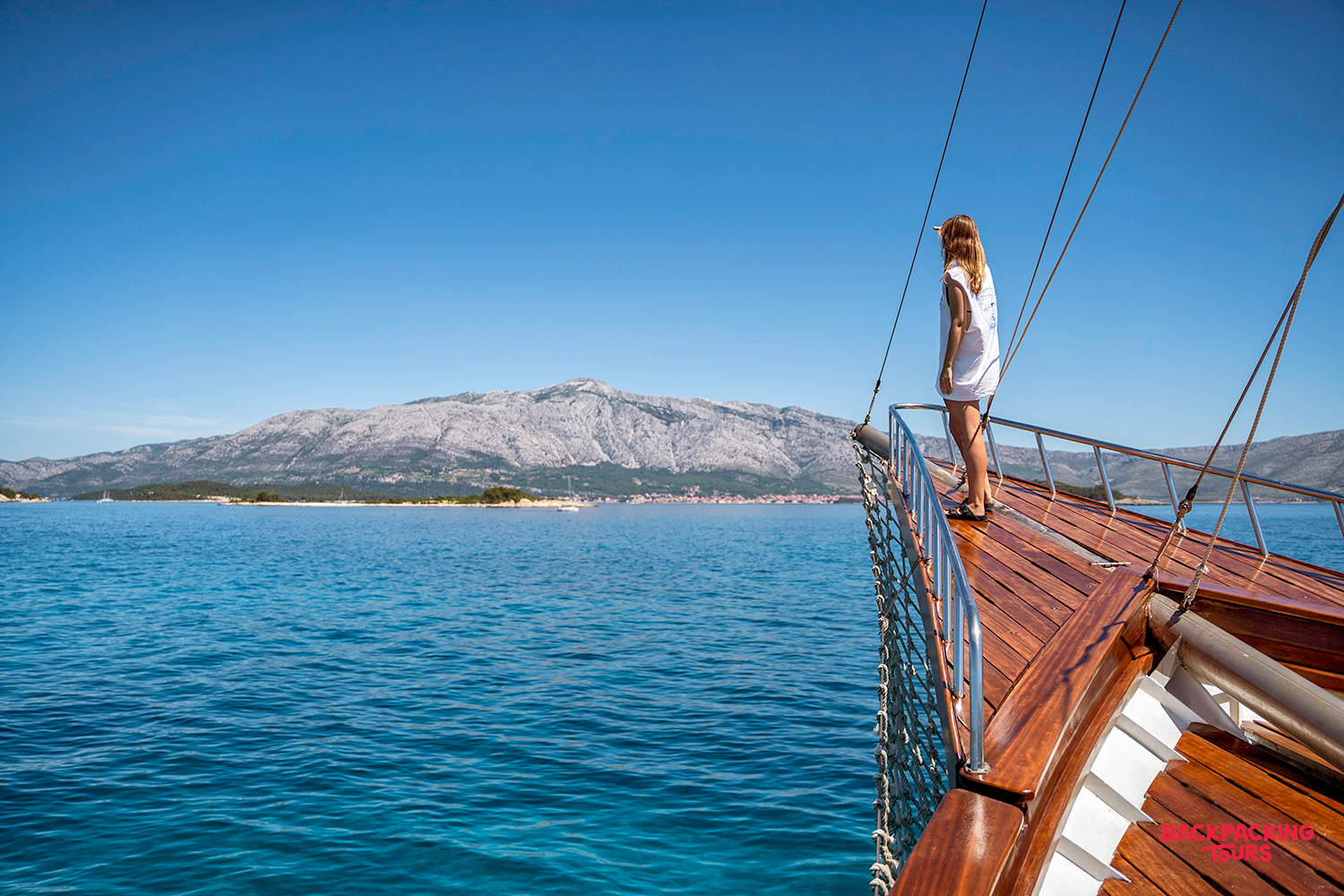 A woman stands at the bow of a sailing boat, admiring the stunning Croatian coastline.
