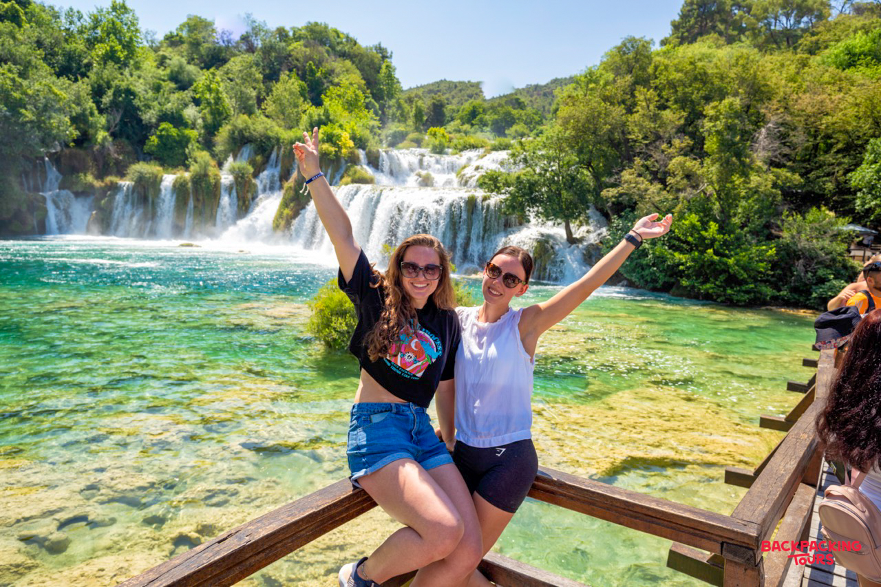 Two happy travelers posing with hands up at the magnificent Krka Waterfalls.