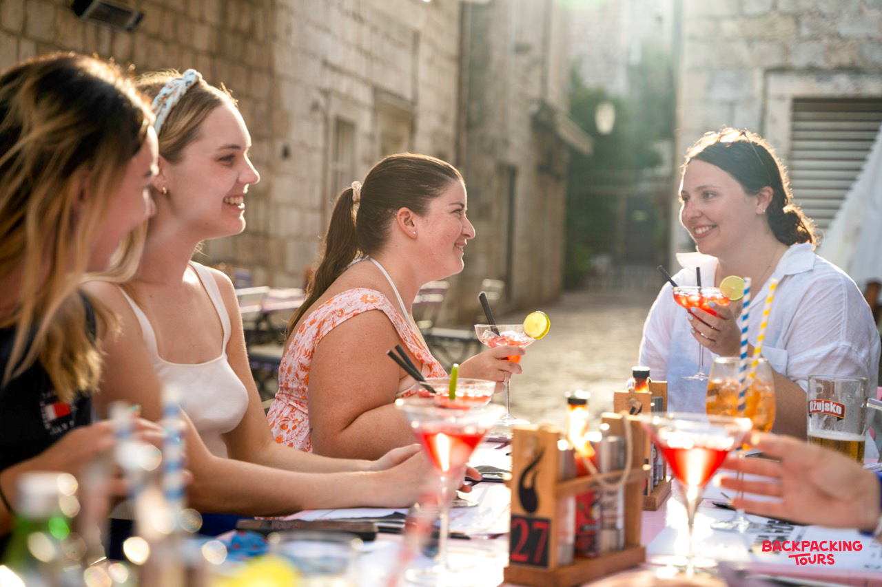 Four happy women enjoy cocktails at an outdoor bar on a sunlit street in Dubrovnik.