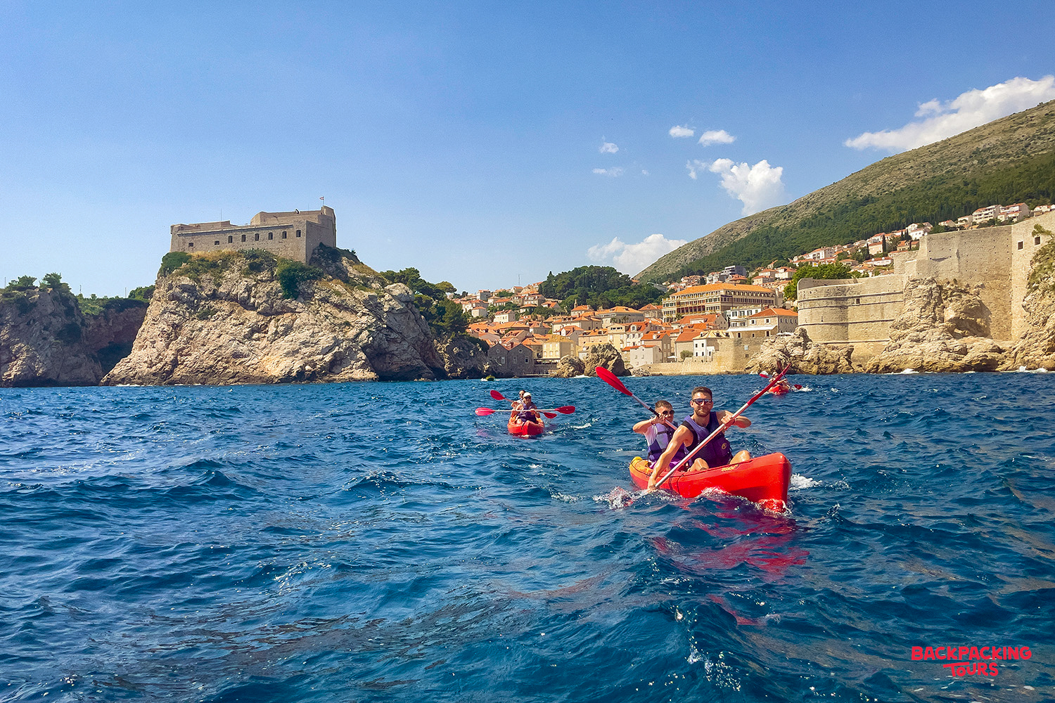 Kayakers paddle on the clear blue Adriatic Sea, passing Dubrovnik's ancient city walls and fortress.